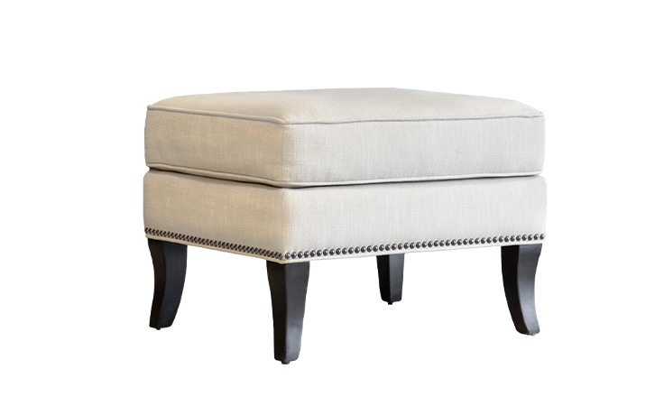 Angled shot of the Atticus Ottoman by Vogel with wooden legs, cream fabric, and nailhead trim
