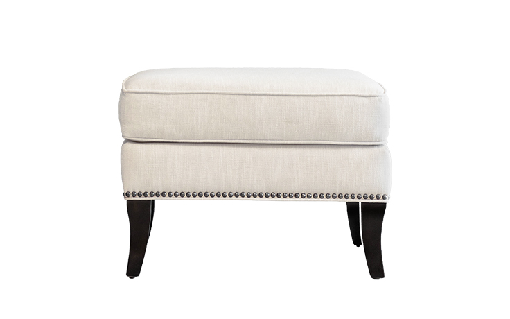 Straight-on shot of the Atticus Ottoman by Vogel with wooden legs, cream fabric, and nailhead trim